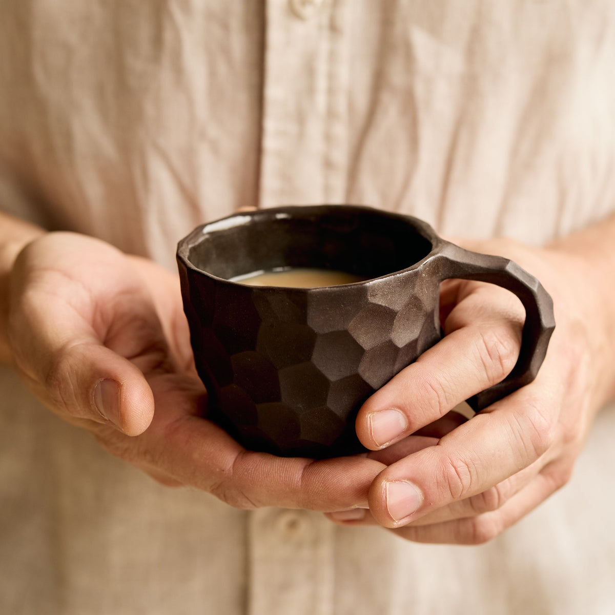 A dark brown carved ceramic cup filled with a milk coffee - person in a beige linen shirt holding it. 