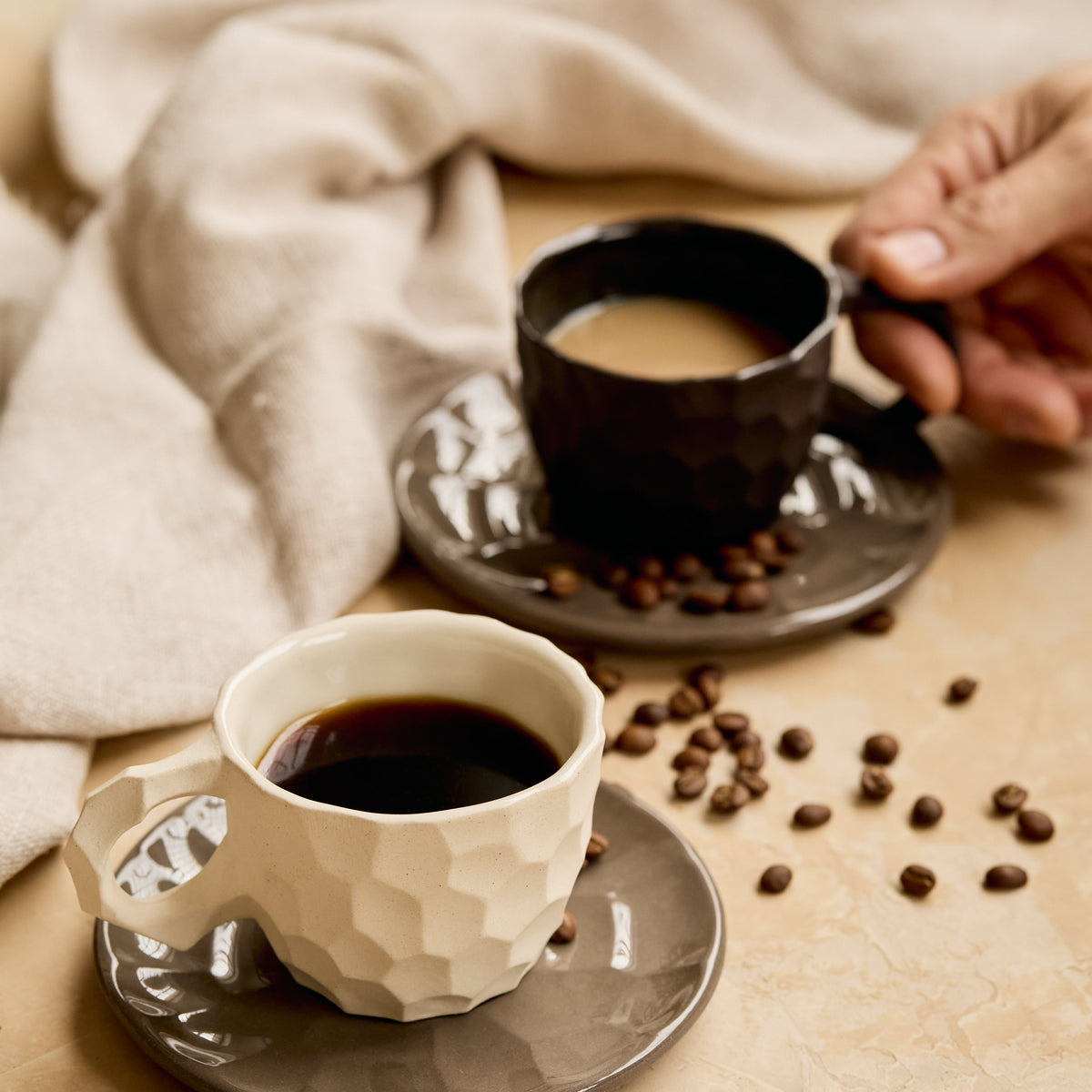 Two textured ceramic coffee cups and saucers on a beige surface with scattered coffee beans and a draped cream cloth. One cup is filled with black coffee in the foreground, and a hand holds the handle of the second cup filled with light coffee in the background.