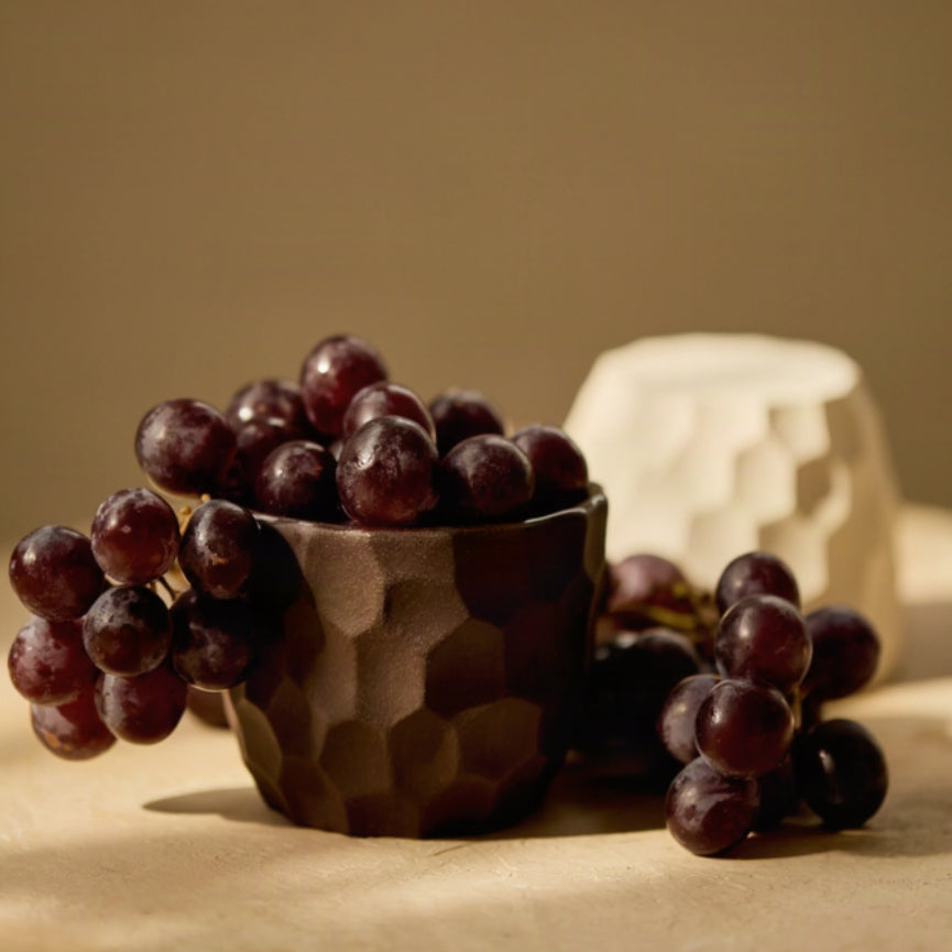 A dark brown textured ceramic cup filled with red grapes, set on a beige surface against a warm brown background, with a white carved cup behind it.