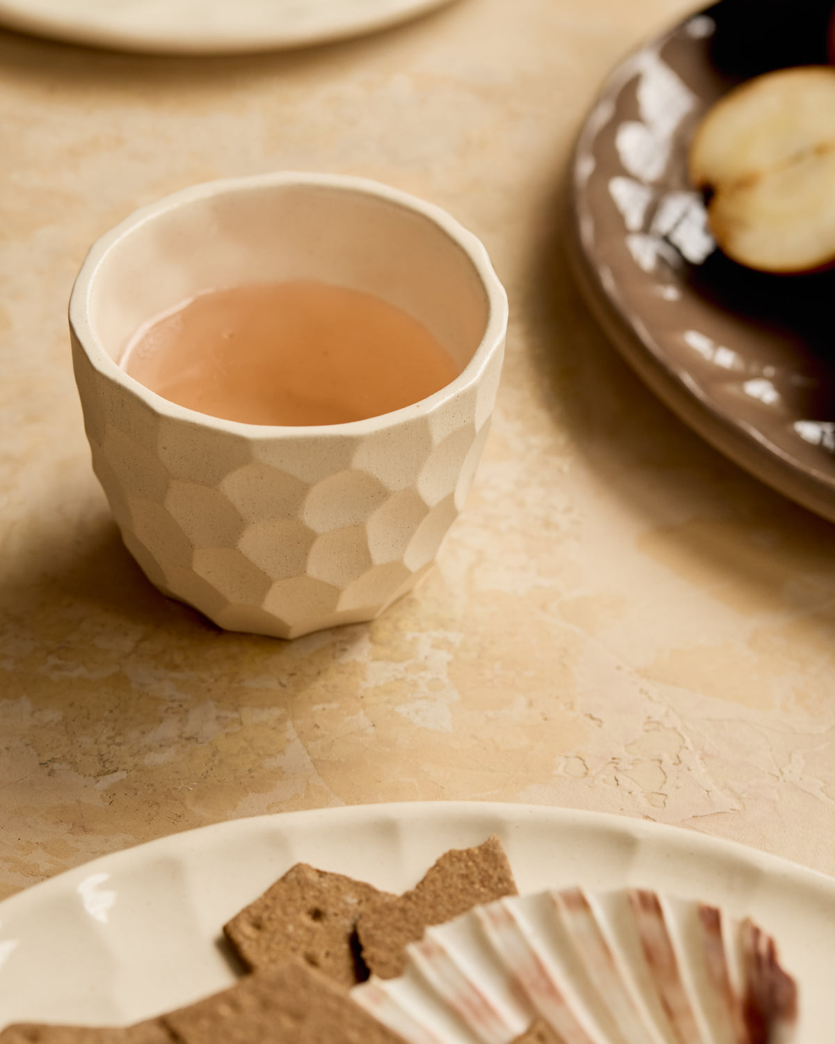 A carved ceramic cup filled with wine drink, placed on a beige stone-like surface with plates and food partially visible in a softly styled setting.